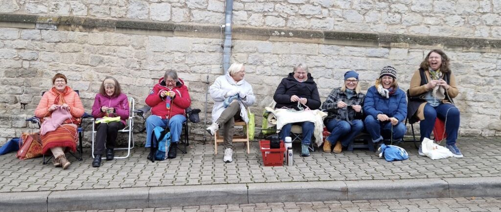 acht Frauen, vor einer Mauer sitzend, handarbeitend